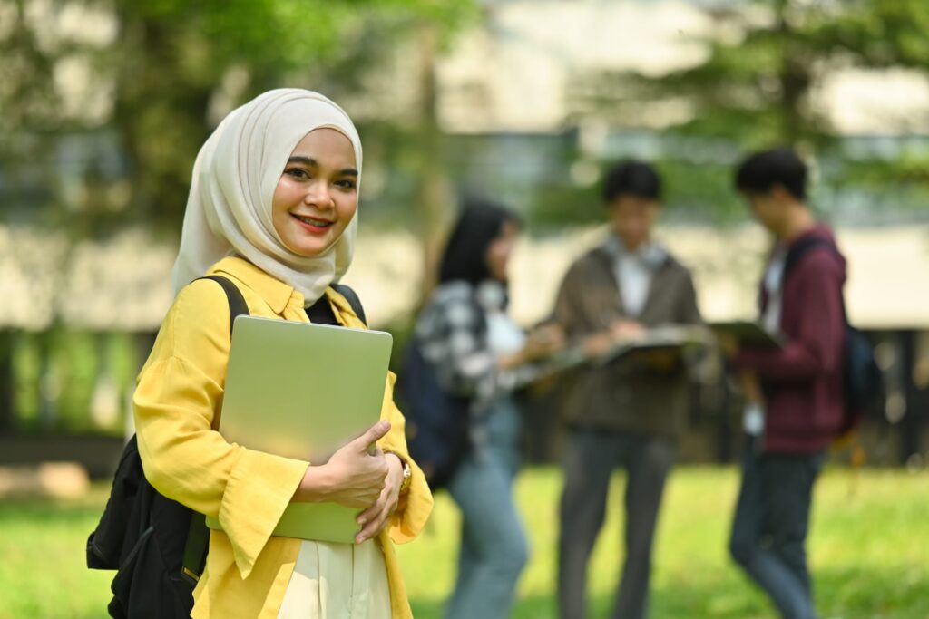 In the bustling environment of university life, this smiling Asian Muslim woman stands confidently with her laptop, embodying determination and aspiration. Surrounded by fellow students, she represents the vibrant diversity that enriches academic spaces.